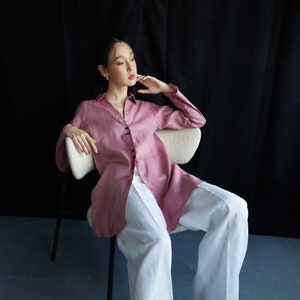 May include: A woman wearing a pink button-down shirt and white wide-leg trousers sits in a chair with a white cushion. She is wearing black shoes.
