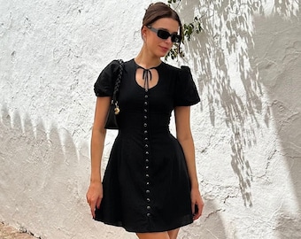 May include: A black mini dress with a button-up front, puff sleeves, and a keyhole neckline. The dress is worn with black strappy heels.