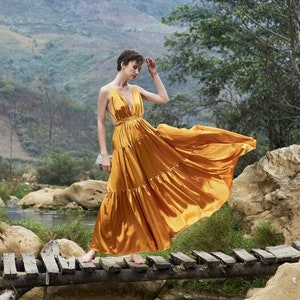 May include: A woman in a flowing gold dress stands on a wooden bridge over a stream. The dress is ruffled and billows in the wind. The background is a lush green landscape with mountains in the distance.