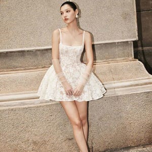May include: A white lace mini dress with a square neckline and thin straps. The dress has a fitted bodice and a flared skirt. The dress is paired with white heels.