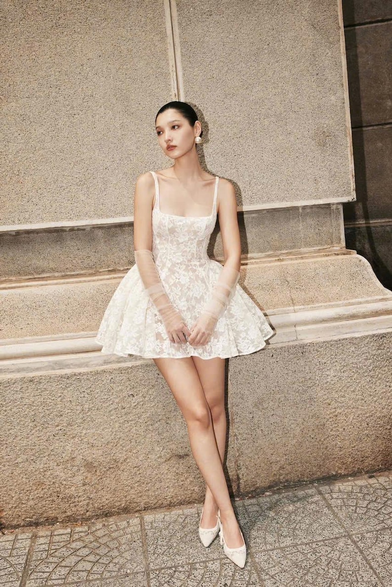 May include: A white lace mini dress with a square neckline and thin straps. The dress has a fitted bodice and a flared skirt. The dress is paired with white heels.