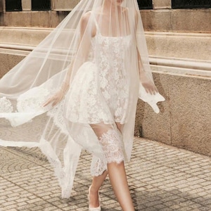 May include: A white lace wedding dress with a long veil. The dress has a fitted bodice and a flowing skirt. The bride is wearing white shoes.