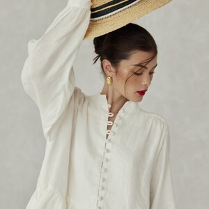 May include: A white linen blouse with a button-up front and a ruffled hem. The blouse has long sleeves and a relaxed fit. The model is wearing a straw hat with a black and white ribbon.