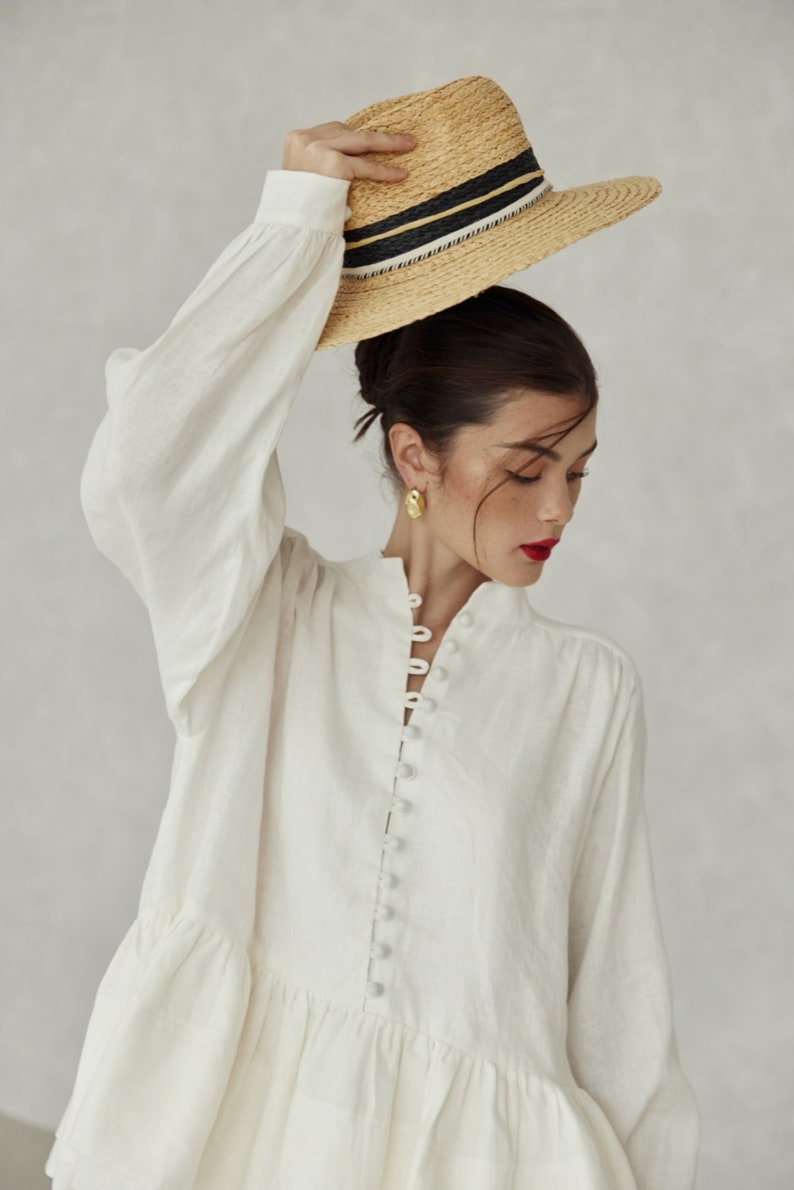 May include: A white linen blouse with a button-up front and a ruffled hem. The blouse has long sleeves and a relaxed fit. The model is wearing a straw hat with a black and white ribbon.