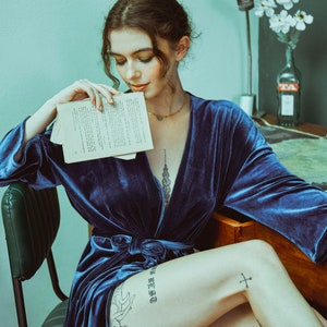 May include: A woman wearing a blue velvet robe sits on a chair, holding a book in her hands. She has a tattoo on her leg that says "D.S.E.S.O." and a cross tattoo on her thigh.
