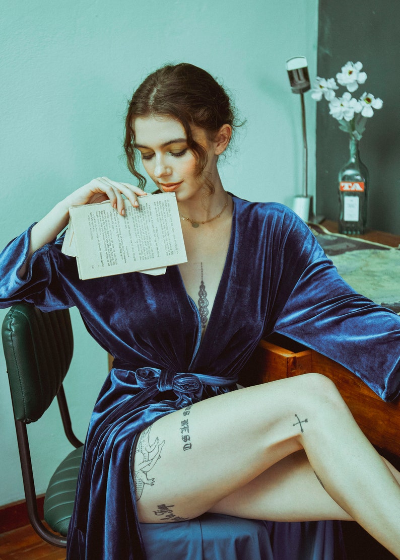 May include: A woman wearing a blue velvet robe sits on a chair, holding a book in her hands. She has a tattoo on her leg that says "D.S.E.S.O." and a cross tattoo on her thigh.