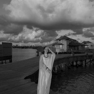 May include: A woman wearing a white dress stands on a wooden walkway over water. The walkway is connected to a building in the background. The sky is cloudy and the water is calm.