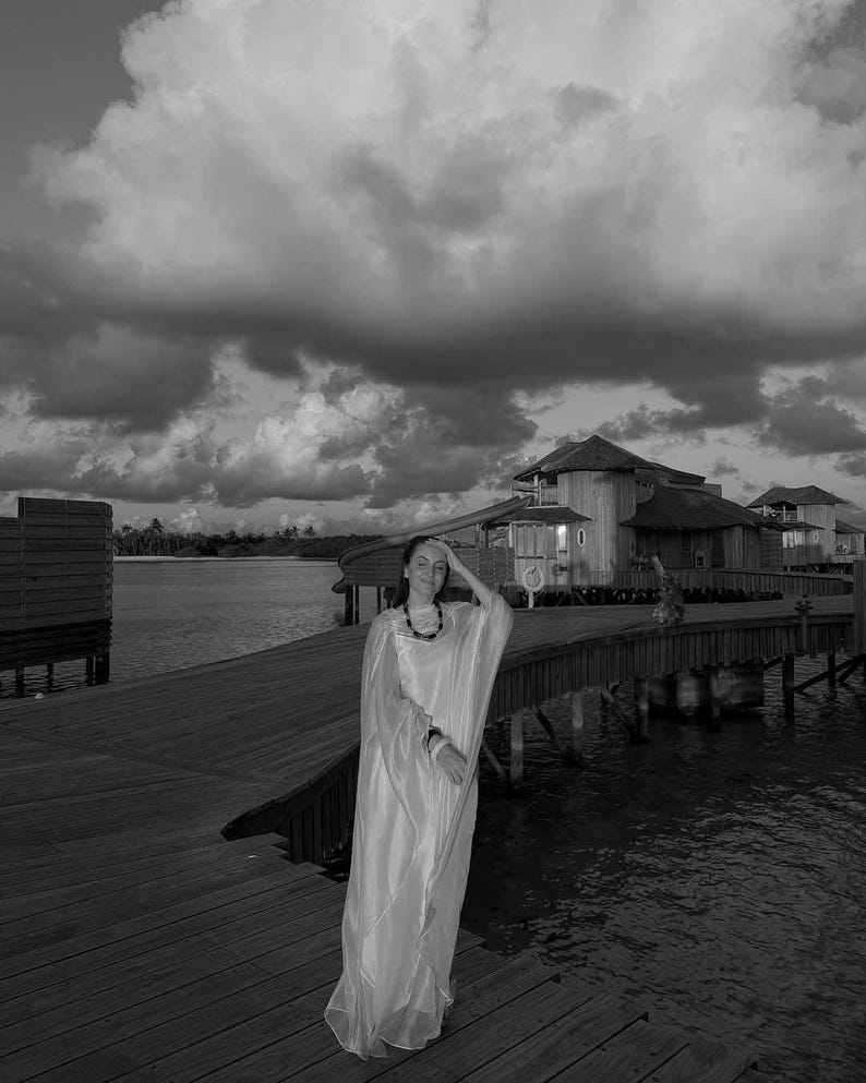 May include: A woman wearing a white dress stands on a wooden walkway over water. The walkway is connected to a building in the background. The sky is cloudy and the water is calm.