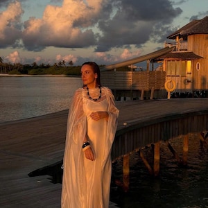 May include: A woman wearing a white dress and a black necklace stands on a wooden dock overlooking a body of water. The sky is a soft orange and pink, and there is a wooden building in the background.