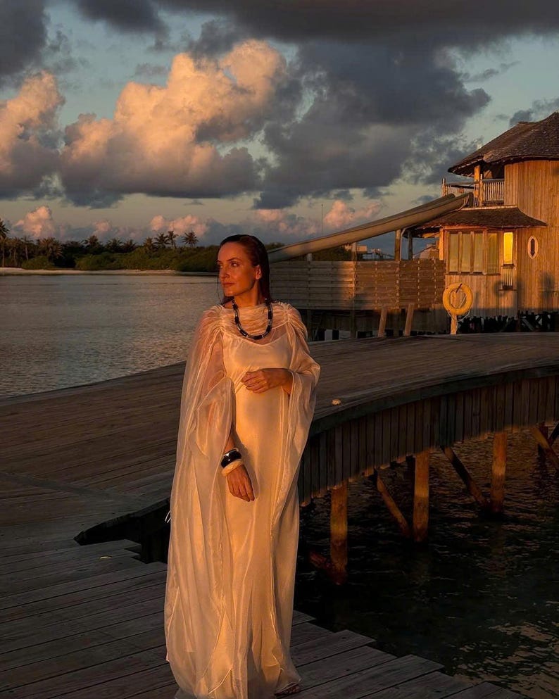 May include: A woman wearing a white dress and a black necklace stands on a wooden dock overlooking a body of water. The sky is a soft orange and pink, and there is a wooden building in the background.