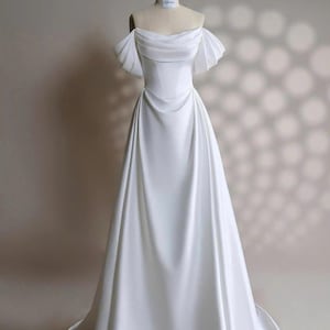 May include: A white off-the-shoulder wedding dress with a fitted bodice and a flowing A-line skirt. The dress has short, ruffled sleeves and a simple, elegant design.