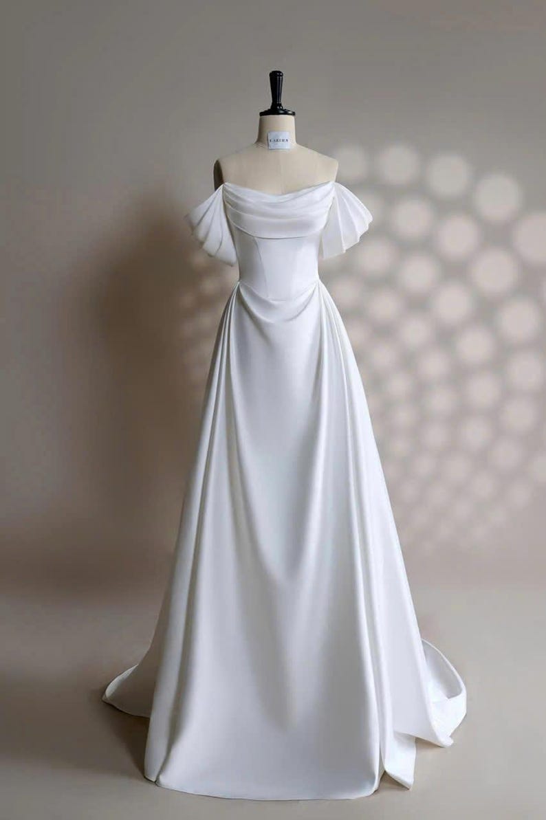May include: A white off-the-shoulder wedding dress with a fitted bodice and a flowing A-line skirt. The dress has short, ruffled sleeves and a simple, elegant design.
