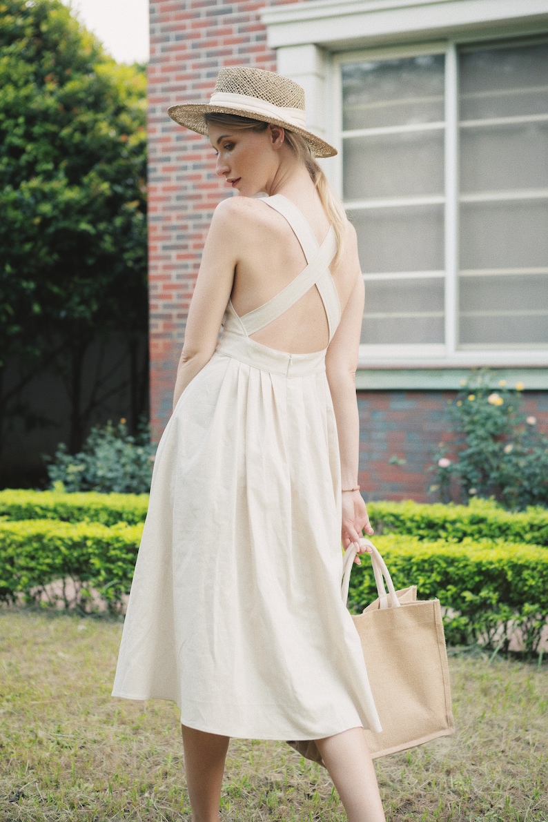 May include: A woman wearing a white linen dress with a criss-cross back detail and a straw hat. She is carrying a beige tote bag.