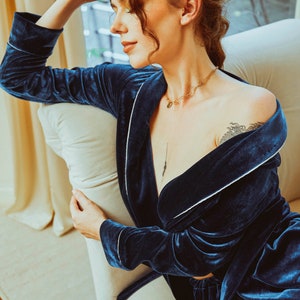K&ouml;nnte beinhalten: Eine Frau tr&auml;gt eine dunkelblaue Samt-Robe mit wei&szlig;en Paspeln. Die Robe ist offen und zeigt eine goldene Kettenkette mit einem Anh&auml;nger. Die Frau sitzt auf einem wei&szlig;en Stuhl mit einem wei&szlig;en Kissen.