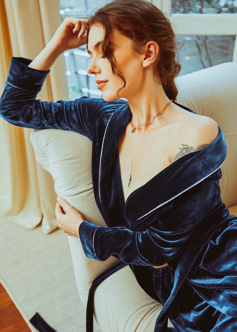 K&ouml;nnte beinhalten: Eine Frau tr&auml;gt eine dunkelblaue Samt-Robe mit wei&szlig;en Paspeln. Die Robe ist offen und zeigt eine goldene Kettenkette mit einem Anh&auml;nger. Die Frau sitzt auf einem wei&szlig;en Stuhl mit einem wei&szlig;en Kissen.