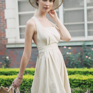 May include: A woman wearing a white linen dress with a straw hat and holding a brown tote bag.