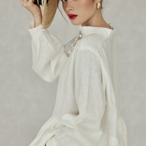 May include: A white linen blouse with a high neckline and long sleeves. The blouse has a ruffled hem and a button closure at the neck. The model is wearing the blouse with a straw hat.