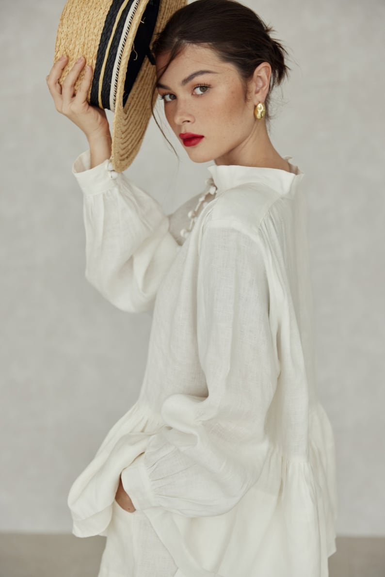 May include: A white linen blouse with a high neckline and long sleeves. The blouse has a ruffled hem and a button closure at the neck. The model is wearing the blouse with a straw hat.
