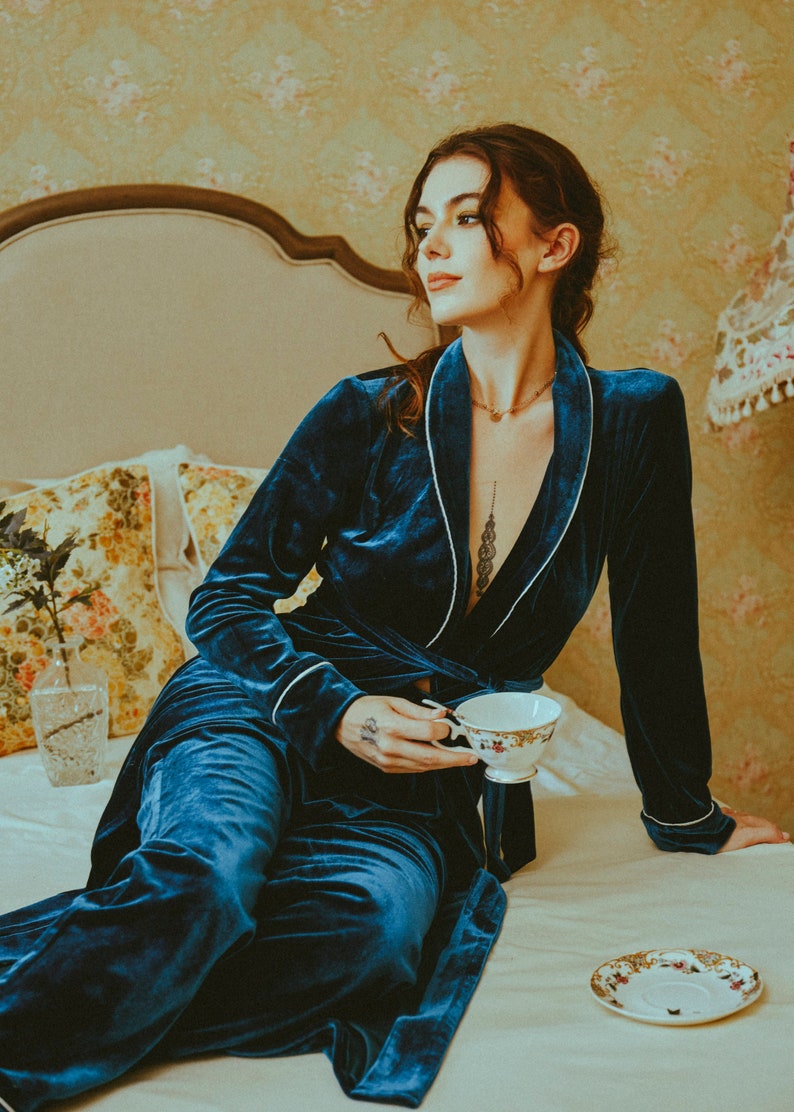 K&ouml;nnte beinhalten: Eine Frau in einem blauen Samt-Morgenmantel mit wei&szlig;en Paspeln sitzt auf einem Bett und h&auml;lt eine Teetasse. Der Morgenmantel hat einen Schalkragen und einen G&uuml;rtel in der Taille. Die Teetasse ist wei&szlig; mit goldenen Verzierungen und floralen Mustern.