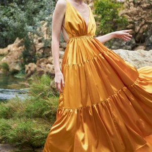 May include: A woman wearing a long, flowing, gold satin dress with a deep V-neckline and a tiered skirt. The dress has a fitted bodice and a loose, flowing skirt. The woman is standing on a wooden platform.