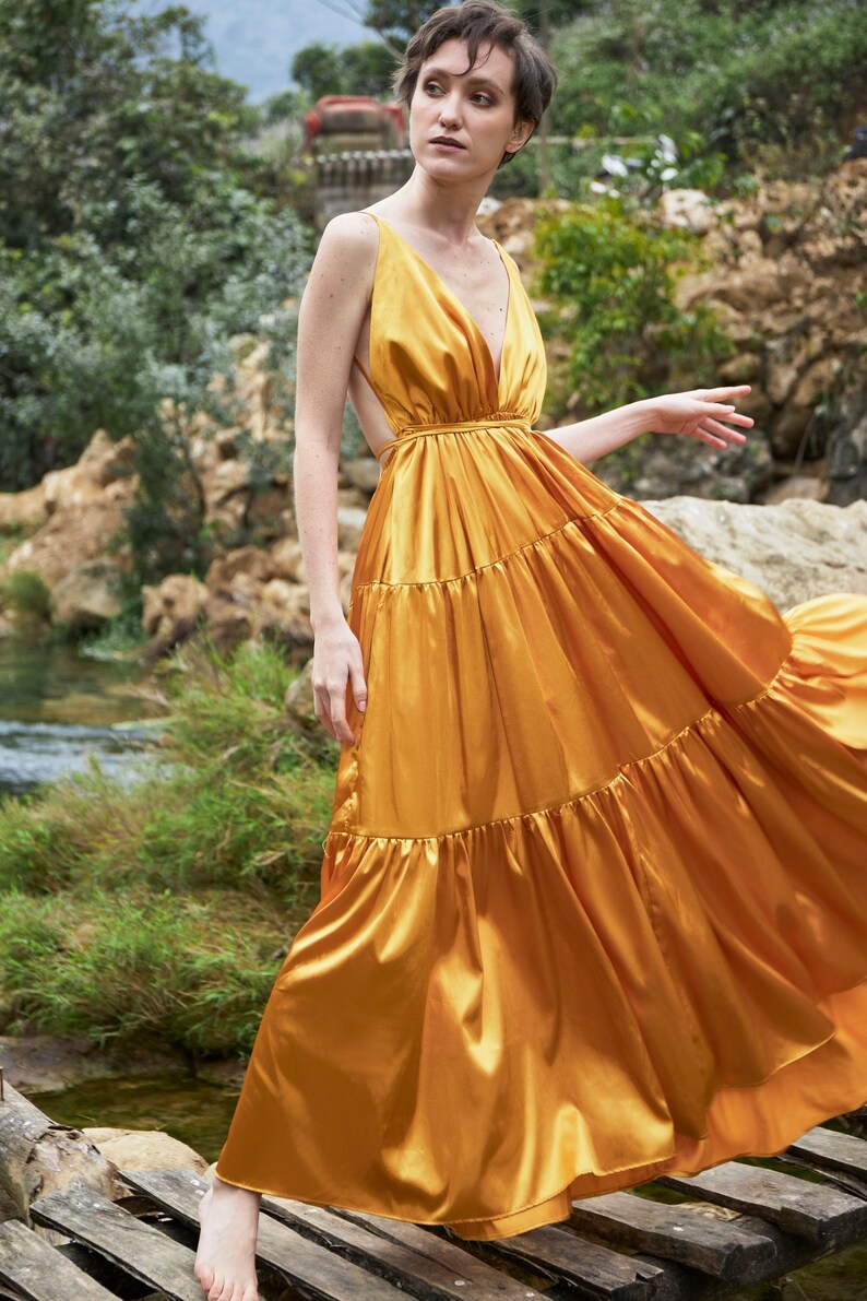 May include: A woman wearing a long, flowing, gold satin dress with a deep V-neckline and a tiered skirt. The dress has a fitted bodice and a loose, flowing skirt. The woman is standing on a wooden platform.