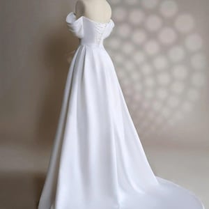 May include: A white off-the-shoulder wedding dress with a fitted bodice and a flowing skirt. The dress has a lace-up back and a train.