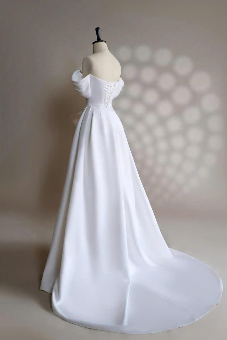 May include: A white off-the-shoulder wedding dress with a fitted bodice and a flowing skirt. The dress has a lace-up back and a train.