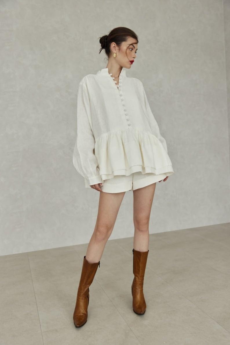 May include: A white linen blouse with a ruffled hem and a button-up front. The blouse is worn with white shorts and brown leather boots.