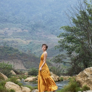 May include: A woman in a gold dress walks across a wooden bridge over a stream in a lush green valley. The mountains in the background are covered in fog.