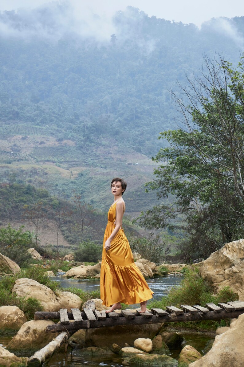 May include: A woman in a gold dress walks across a wooden bridge over a stream in a lush green valley. The mountains in the background are covered in fog.