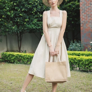 May include: A woman wearing a straw hat and a white linen dress holds a brown tote bag. She is standing in a grassy area with green bushes behind her.