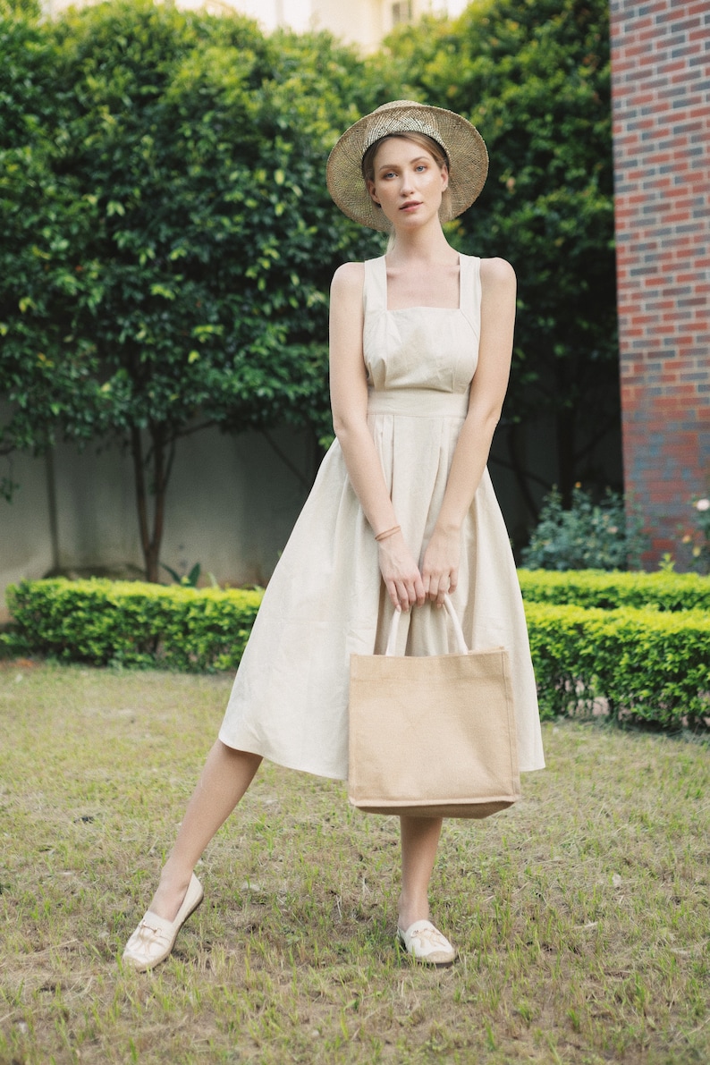 May include: A woman wearing a straw hat and a white linen dress holds a brown tote bag. She is standing in a grassy area with green bushes behind her.