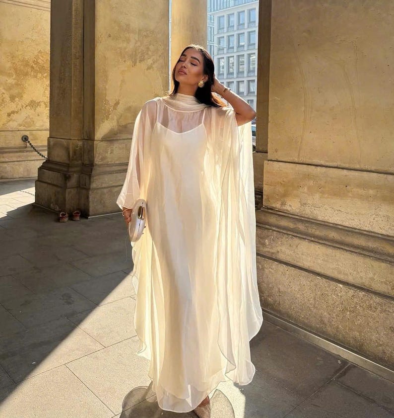 May include: A white sheer dress with a long, flowing skirt and a high neckline. The dress has a simple design and is made of a lightweight fabric.