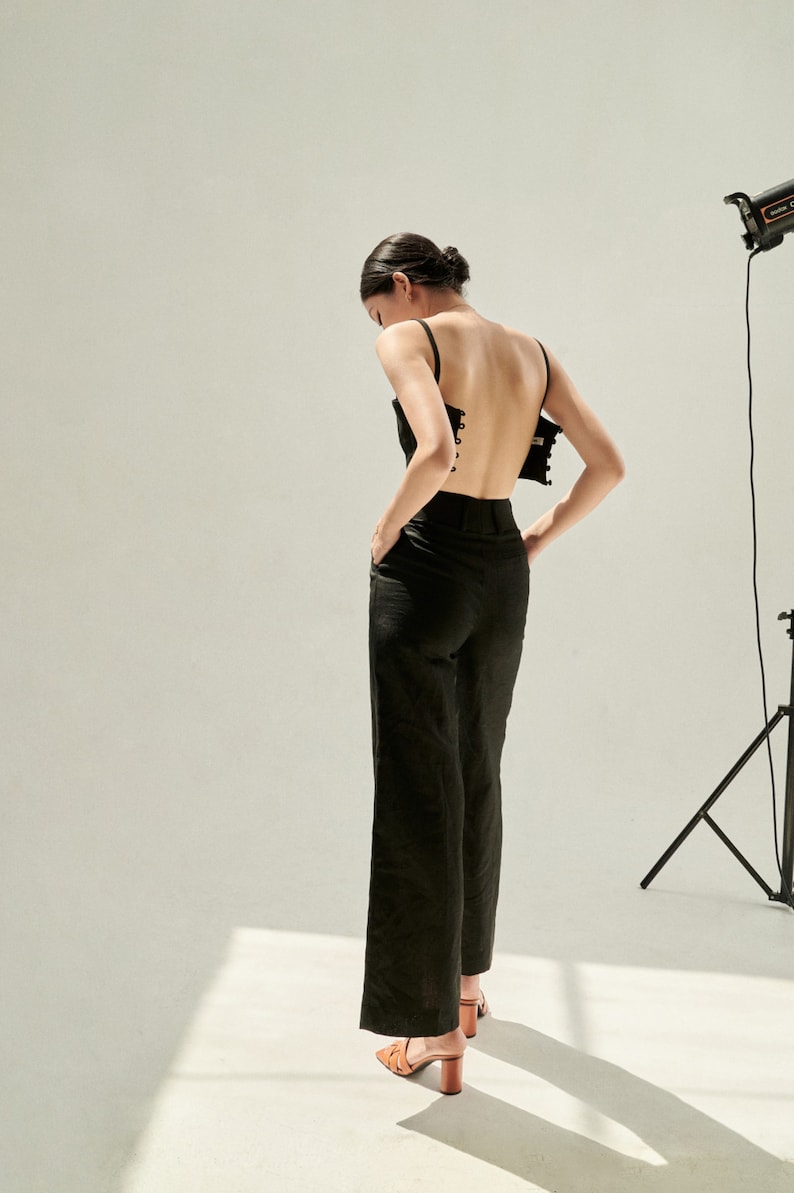 K&ouml;nnte beinhalten: Eine Frau tr&auml;gt ein schwarzes, r&uuml;ckenfreies Oberteil mit Schn&uuml;rung und eine schwarze, weite Hose. Sie steht in einem Studio mit wei&szlig;em Hintergrund.