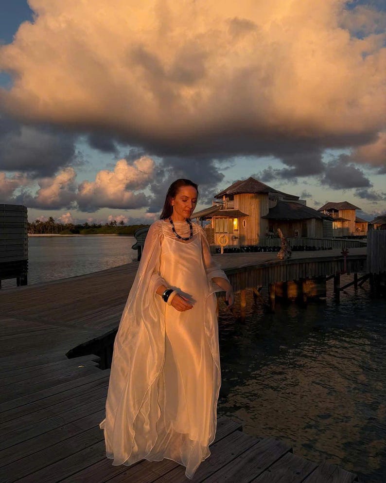 May include: A woman wearing a white flowing dress and a dark necklace stands on a wooden dock over the water. The sky is a soft orange and pink with large clouds. The background shows a wooden building with a thatched roof.