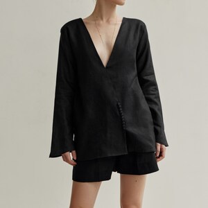 May include: A black linen shirt with a deep V-neckline and a button detail on the side. The shirt is worn with black shorts.