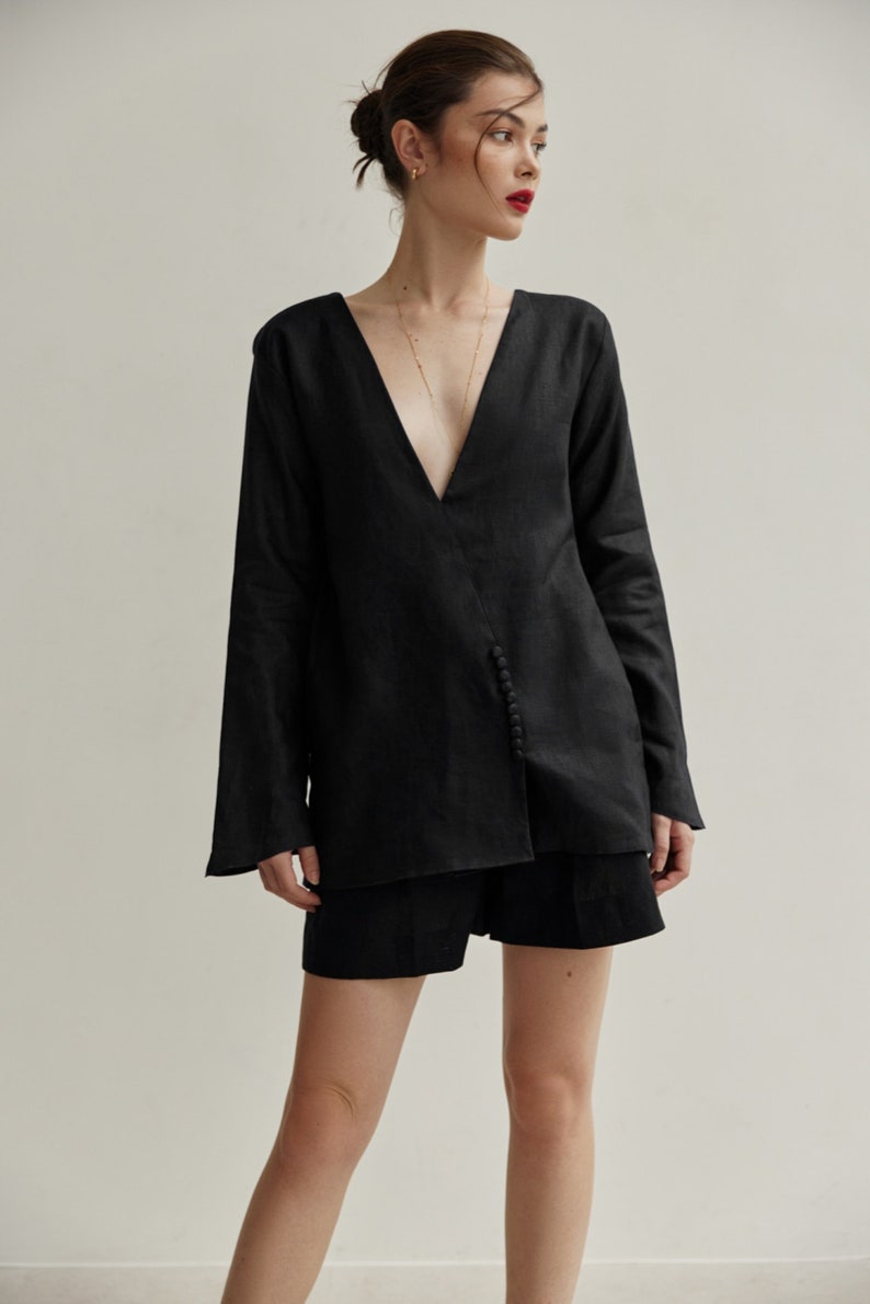 May include: A black linen shirt with a deep V-neckline and a button detail on the side. The shirt is worn with black shorts.