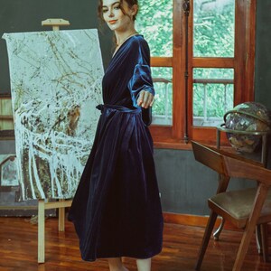May include: A woman wearing a dark blue velvet robe with a sash tie. She is standing in a room with a wooden floor and a painting on an easel.