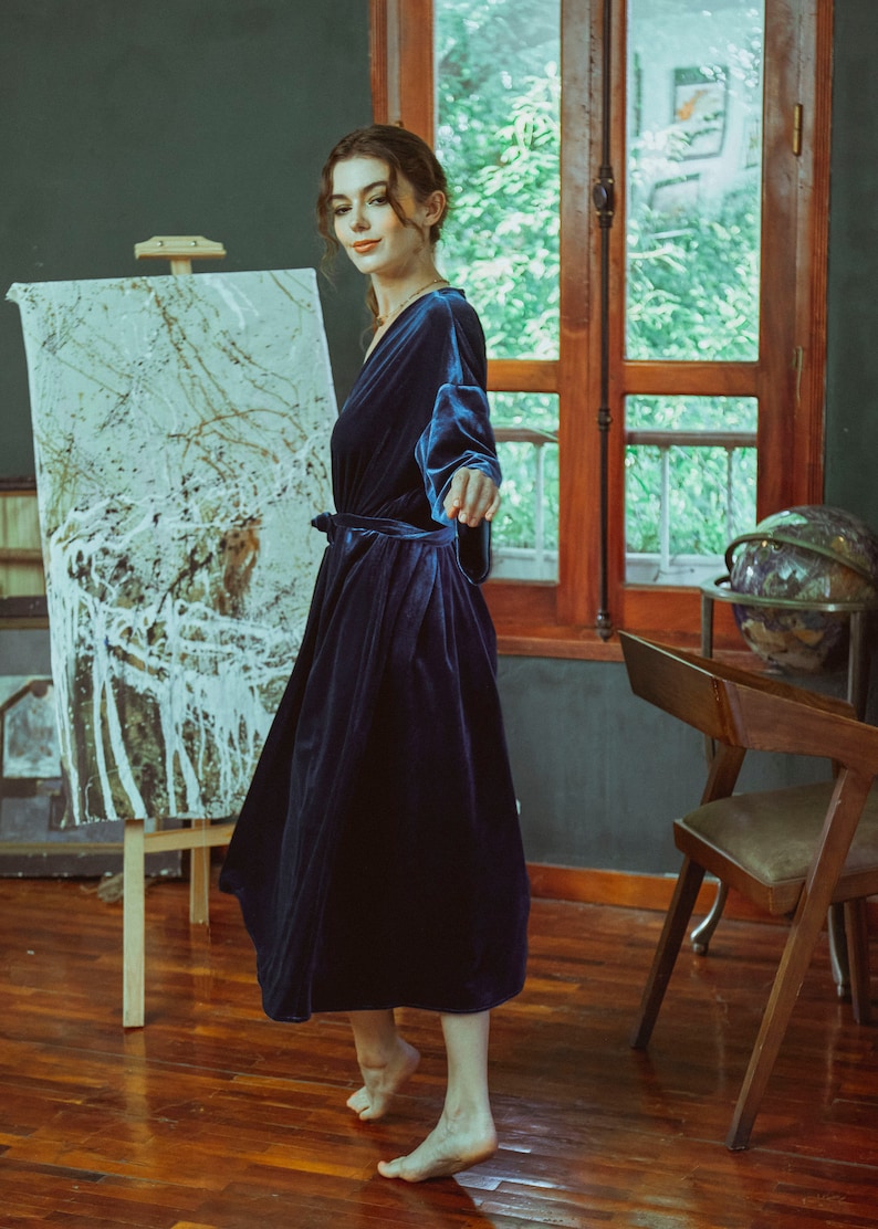 May include: A woman wearing a dark blue velvet robe with a sash tie. She is standing in a room with a wooden floor and a painting on an easel.