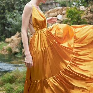 May include: A woman wearing a long, flowing, yellow satin dress with a low-cut neckline and a tiered skirt. The dress has a cutout back and is cinched at the waist.