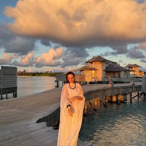 May include: A woman wearing a white dress stands on a wooden walkway over the water. The walkway is surrounded by water and wooden buildings. The sky is a light orange color with white clouds.