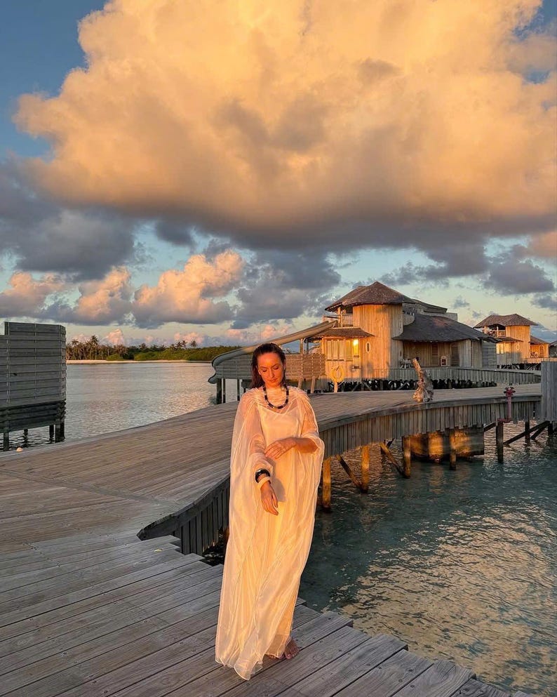 May include: A woman wearing a white dress stands on a wooden walkway over the water. The walkway is surrounded by water and wooden buildings. The sky is a light orange color with white clouds.