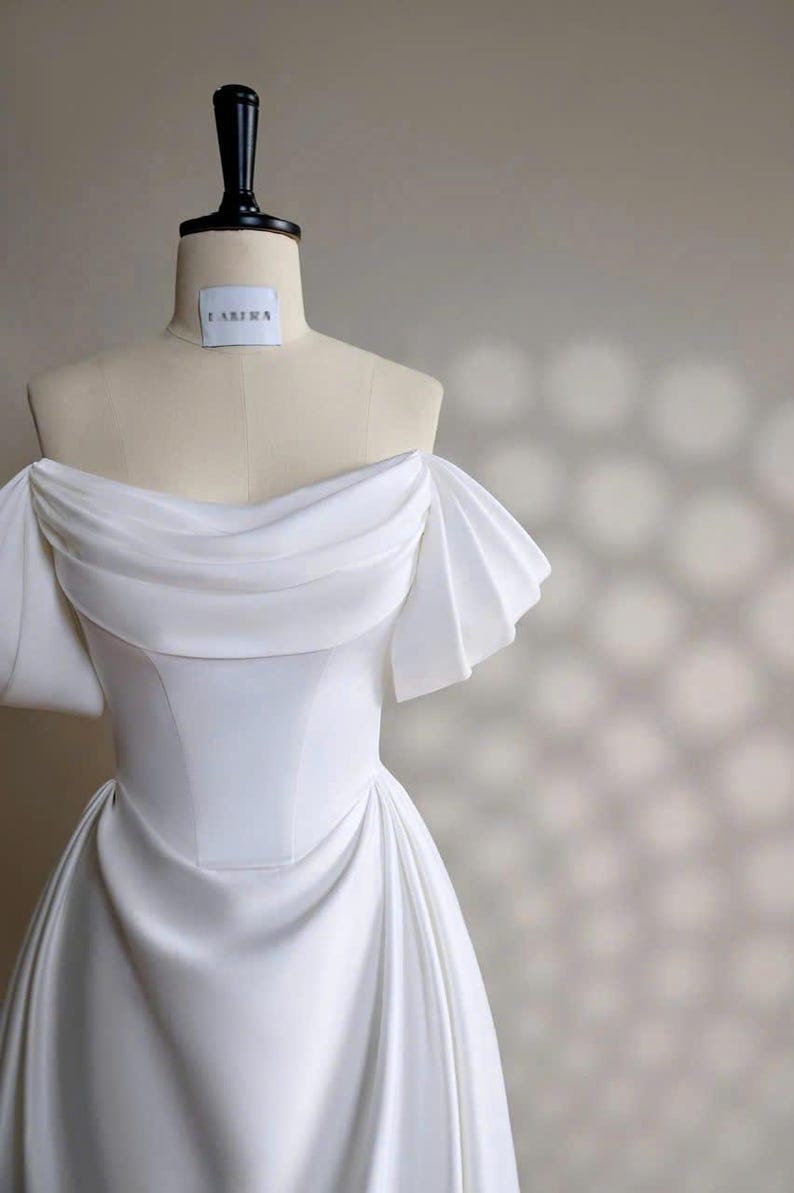 May include: A white off-the-shoulder wedding dress with a fitted bodice and a flowing skirt. The dress has a simple design with a draped neckline and short, pleated sleeves. The dress is displayed on a mannequin against a light beige background.