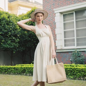 May include: A woman wearing a white dress and a straw hat is holding a beige tote bag. She is standing in a grassy area with a brick building in the background.