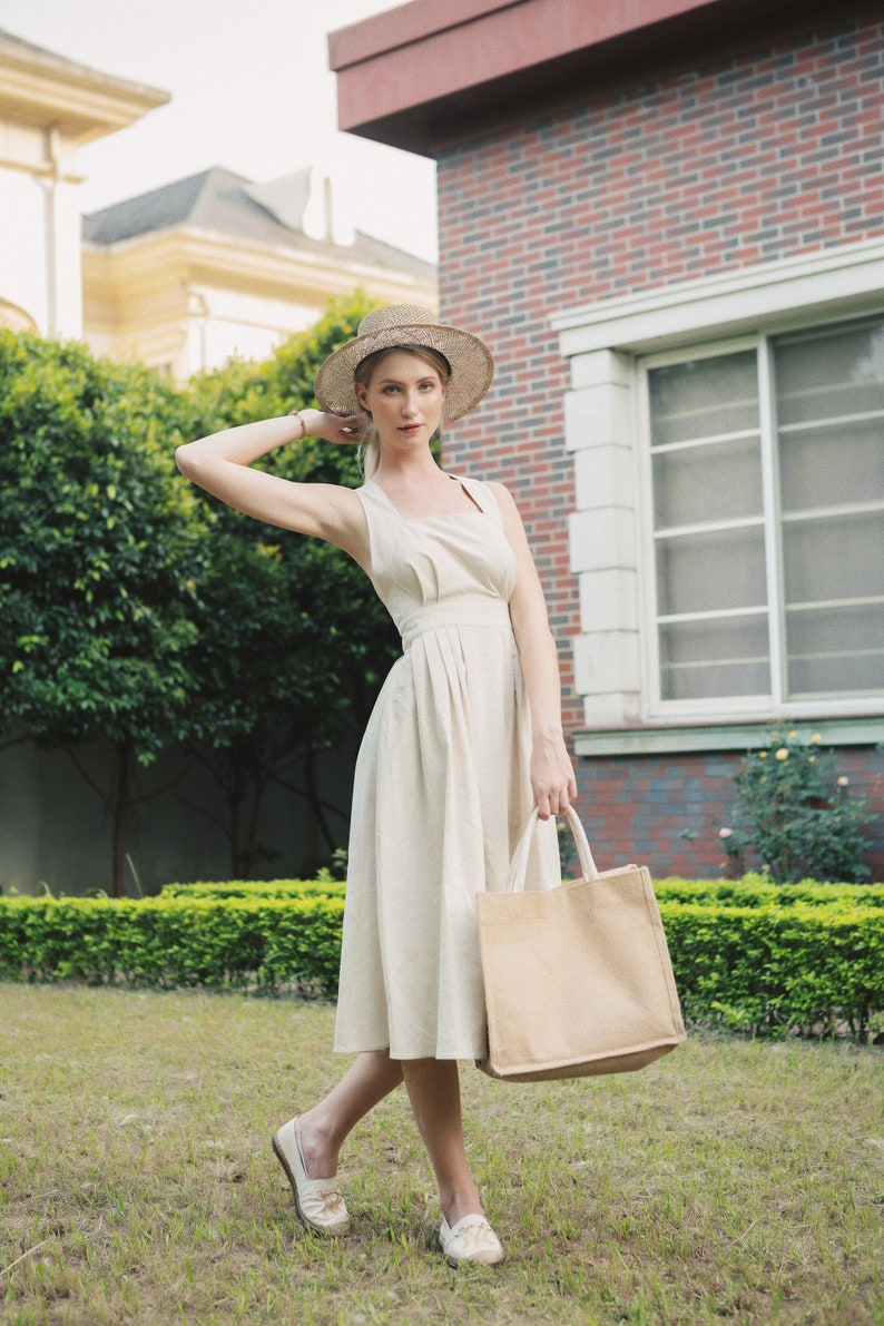 May include: A woman wearing a white dress and a straw hat is holding a beige tote bag. She is standing in a grassy area with a brick building in the background.