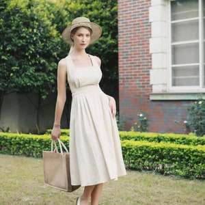 May include: A woman wearing a white sundress and a straw hat holds a brown tote bag. She is standing in a grassy area with a brick building in the background.