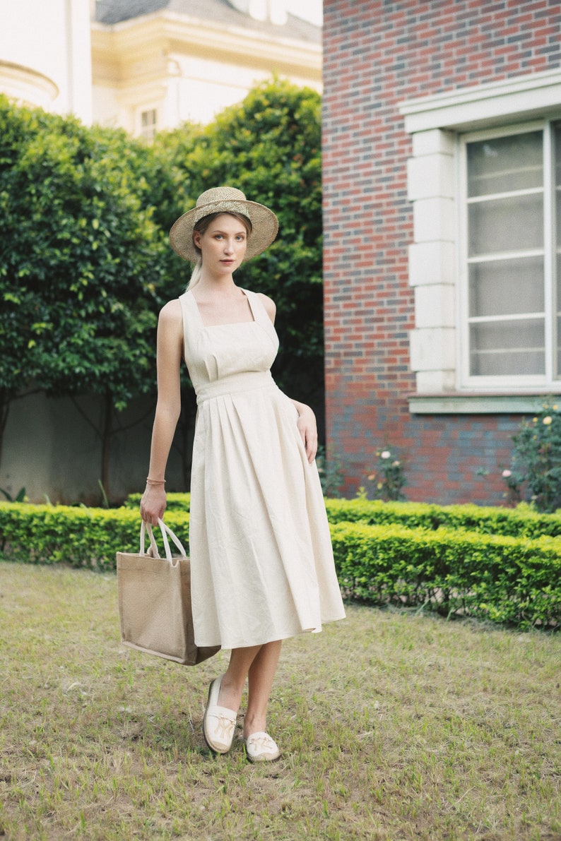 May include: A woman wearing a white sundress and a straw hat holds a brown tote bag. She is standing in a grassy area with a brick building in the background.