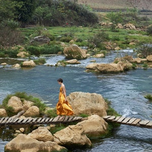 May include: A person in a yellow dress walks across a wooden bridge over a stream. The stream is surrounded by rocks and green vegetation.