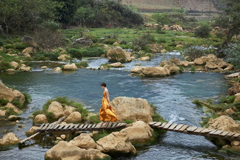 May include: A person in a yellow dress walks across a wooden bridge over a stream. The stream is surrounded by rocks and green vegetation.