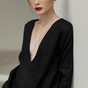 May include: A woman wearing a black, long-sleeved, v-neck top with a gold chain necklace. The top has a button closure and the woman is standing against a white wall.